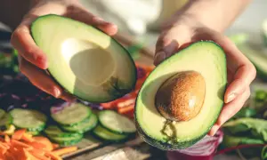 Manos sostienen un aguacate cortado por la mitad, listo para disfrutar. Es la temporada del aguacate en España, con verduras frescas de fondo.