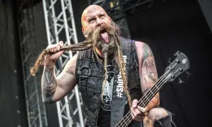 Chris Kael, basista z długą, zaplecioną brodą, wyciąga język podczas koncertu, trzymając gitarę basową.