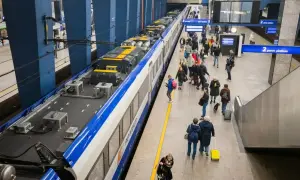 Ludzie z bagażem czekają na peronie PKP. Widać pociąg, tablice informacyjne i schody ruchome.