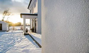 Nowe tynkowanie ścian zewnętrznych domu w zimowej scenerii. Słońce przebija się przez chmury, oświetlając śnieżny krajobraz i białą elewację.