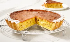 Torta di mele con crema pasticcera, con una fetta tagliata, cosparsa di zucchero a velo su una griglia metallica.