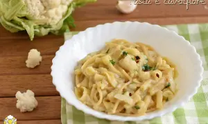 Un piatto di pasta e cavolfiore cremoso, guarnito con prezzemolo e peperoncino. Sullo sfondo, un cavolfiore fresco e uno spicchio d'aglio.