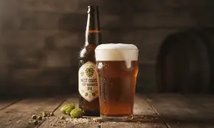 Boccale di birra IPA schiumosa accanto a una bottiglia di West Coast Hop Harvest IPA, con luppolo e chicchi sparsi su un tavolo di legno.