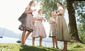 Vier Frauen in Dirndln stoßen am See an. Ein idyllisches Bild für Waldfeste in Bayern.