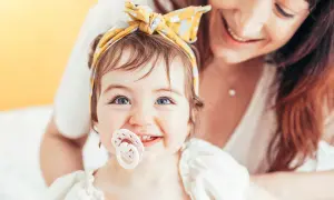 A baby with bright blue eyes and a yellow headband smiles, her passy teeth peeking out from behind a pacifier. Her mother smiles in the background.