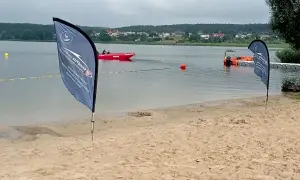 Plaża nad jeziorem, czerwona łódź ratownicza WOPR, pomarańczowa łódź, flagi, budynki na horyzoncie.