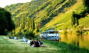 Entspannte Menschen genießen die Sonne am Ufer der Mosel. Ein Ausflugsschiff liegt vor, im Hintergrund steile Weinberge. Perfekte Mosel Ausflugsziele.