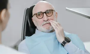 Starszy mężczyzna w okularach siedzi na fotelu dentystycznym, trzymając się za policzek. Wygląda na to, że boli go rozchwiany ząb.