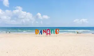 Kolorowe litery "DANANG" na piaszczystej plaży w Wietnamie, z błękitnym morzem i niebem w tle.