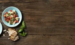 Två tallrikar med tomatsallad och vispad mozzarella, garnerad med basilika. Körsbärstomater och bröd på träbord.
