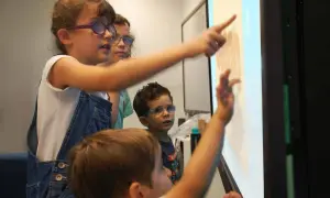 Kinder mit Brillen lernen interaktiv an einem Touchscreen. Sie zeigen auf Zahlen und Symbole, was multisensorisches Lernen ermöglicht.