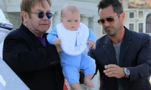 Elton John z dziećmi, w tym małym dzieckiem w niebieskim ubranku i śliniaczku z łódką, w otoczeniu innych mężczyzn.
