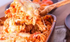 Un cucchiaio solleva una porzione generosa di pasta alla sorrentina, filante e dorata, da una pirofila marrone.