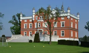 Pałac w Szałszy, ceglana budowla z białymi zdobieniami, otoczona zielonym trawnikiem i drzewami. Na trawniku widać bramkę do piłki nożnej i kilka piłek.