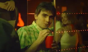Młody mężczyzna w zielonej koszuli pije z czerwonego kubka. W tle widać uśmiechniętą dziewczynę.