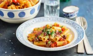 En tallrik med rigatoni pasta med salami, oliver och tomater, toppad med färsk persilja. En skål med mer pasta syns i bakgrunden.