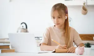 Dziewczynka z słuchawkami uczy się przy laptopie, notując w zeszycie. To nowoczesna nauka w chmurze, która pozwala na elastyczne zdobywanie wiedzy.