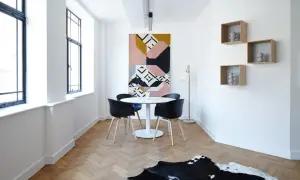 Un comedor moderno con mesa redonda, sillas negras y una alfombra geométrica. Las estanterías flotantes y las ventanas grandes dan la sensación de casas pequeñas y acogedoras.