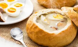 Żurek w chlebie z jajkiem i kiełbasą, obok talerz z jajkami i kiełbasą.