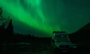 Ein Camper steht unter dem faszinierenden Licht der Aurora Borealis. Grüne Schleier tanzen am sternenklaren Himmel über dunklen Bäumen und einer Bergflanke.