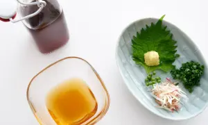 Jaki sos wariacie! Na talerzyku zielona shiso, starty imbir, szczypiorek i zielone pieprzne kuleczki. Obok miseczka z płynnym złotem i butelka z ciemnym płynem.