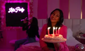 Urodziny Barbie w różowym pokoju. Uśmiechnięta dziewczyna trzyma tort ze świeczkami, a w tle koleżanka pije drinka.