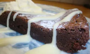 En bit mörk kladdkaka med pudersocker och vit chokladsås, serverad med vispad grädde.