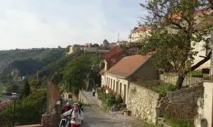 Radfahrer erkunden eine Kopfsteinpflasterstraße in Znaim, Tschechien, mit historischen Gebäuden und einer Burgruine im Hintergrund.