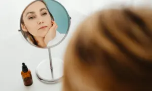 Kobieta w lustrze zastanawia się, czy jest "pick me girl", dotykając policzka. Obok buteleczka z serum.