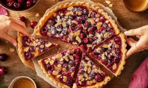 Une délicieuse tarte cerise amande, saupoudrée de sucre glace et d'amandes effilées, est coupée en parts. Des mains tendent pour en prendre une.