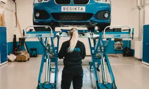 En bil lyfts upp på en lyft för sista besiktningsdag. En mekaniker med flätat hår står framför bilen.