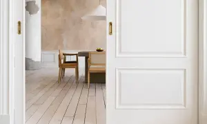 Puertas blancas y suelo de madera clara se combinan para crear un espacio luminoso. Al fondo, un comedor con sillas de madera y una mesa de hormigón.