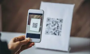 Dłoń trzyma smartfon skanujący kod QR na białej kartce.