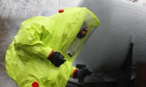 Person in a bright yellow hazmat suit with a clear visor, standing next to a large grey tank.