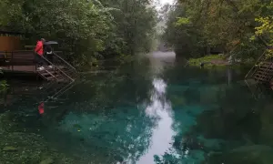 Turkusowe jezioro w Rosji, otoczone drzewami. Dwie osoby stoją na schodach prowadzących do wody, jedna z parasolem.