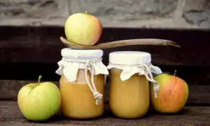 Deux pots de compote de pommes faits maison, décorés de tissus et de cordes, avec des pommes fraîches et une cuillère en bois.