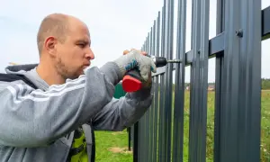 Mężczyzna w rękawiczkach przykręca wkrętarką element do nowego, szarego płotu.