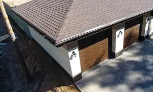 Nowy dach z gontu pokrywa budynek z dwoma brązowymi bramami garażowymi.