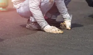 Pracownicy w brudnych rękawicach i jeansach wykonują naprawę dachu z papy, klęcząc na ciemnej powierzchni.