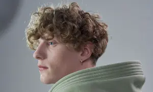 Primo piano di un giovane con capelli mossi uomo, occhi azzurri e un orecchino a cerchio. Indossa una felpa verde chiaro.