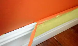Ta bort maskeringstejp efter målning ger en ren kant. Orange vägg möter vit golvlist med gul tejp.