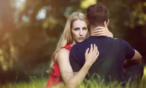 Kobieta w czerwonej sukience obejmuje mężczyznę. Jej spojrzenie wyraża radykalną szczerość, gdy wtula się w niego w trawie.