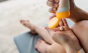 Manos aplicando crema solar caduca en la palma. Un niño descansa en una tumbona al fondo.