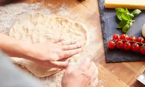 Hände formen den **pizzateig auftauen** auf einer bemehlten Holzoberfläche. Daneben liegen frische Tomaten, Mozzarella und Basilikum.