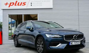 Ciemnoniebieskie Volvo V60 Hybrid z polskimi tablicami WL 7253Y przed budynkiem z logo "+plus".