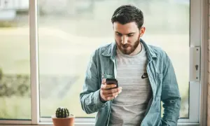 Hombre con barba mira su móvil, quizás buscando frases cuando no te responden. Un cactus en maceta decora el alféizar.