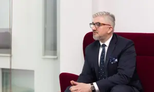 Mężczyzna w garniturze i okularach siedzi na czerwonym fotelu, patrząc w bok.