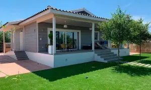 Moderna casa Canexel con porche, césped artificial y árboles frondosos bajo un cielo azul.