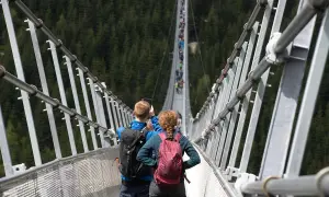 Para idzie przez wiszący most w Czechach, robiąc zdjęcia. W tle widać las i innych turystów.