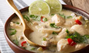 Aksamitna zupa tom kha gai z kawałkami kurczaka, grzybami i chili, udekorowana plasterkami limonki i świeżą kolendrą. Drewniana łyżka zanurzona w aromatycznym bulionie.